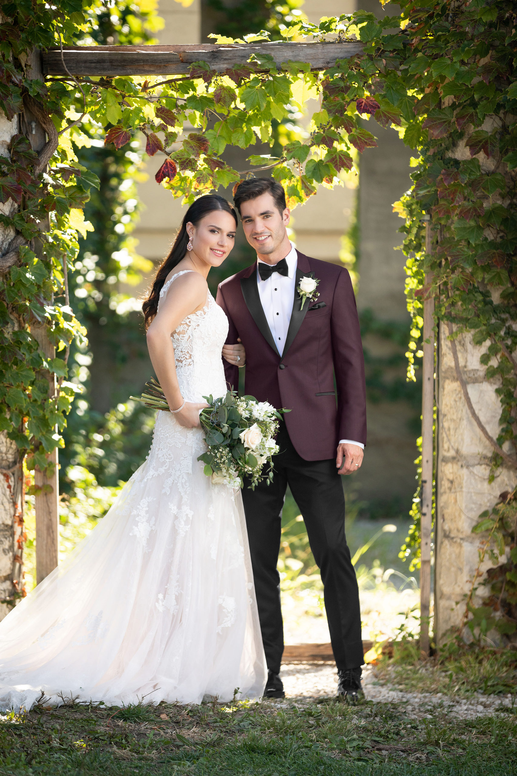 A wedding couple in elegant wedding suits, standing together outdoors. Green leaves create a beautiful natural backdrop